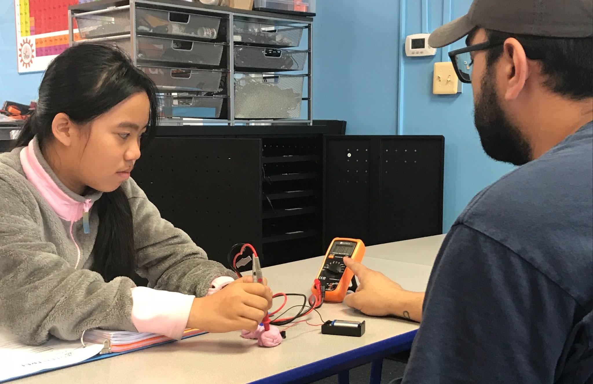 Student receiving one-on-one guidance during a hands-on STEM learning activity using electronic testing equipment.